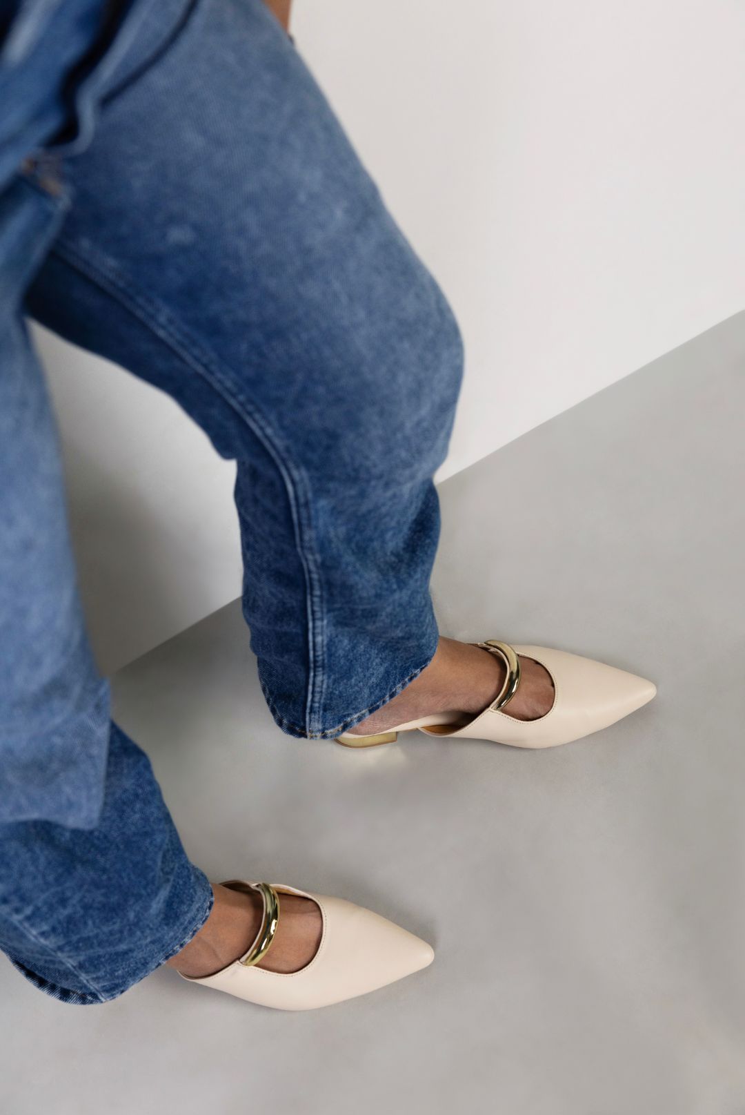 Person wearing blue jeans and beige pointed-toe shoes on a light gray floor.