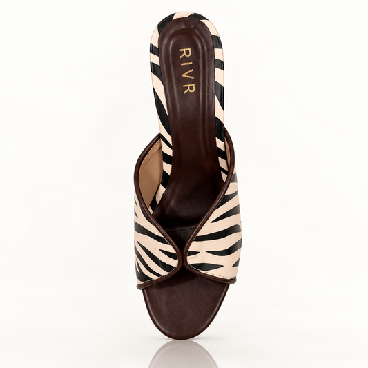 A single women's heeled slide sandal by RIVR, featuring a zebra-print upper in beige and black with a dark brown leather toe strap and heel counter, set against a plain white background with a soft reflection below.